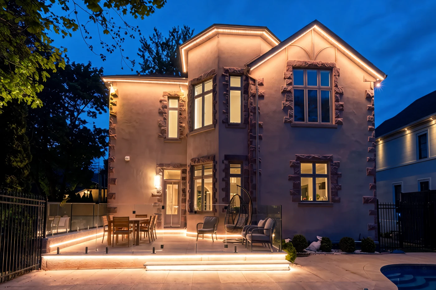 Real stone home at dusk with warm-white light bars tracing the roofline and facade, lit patio steps